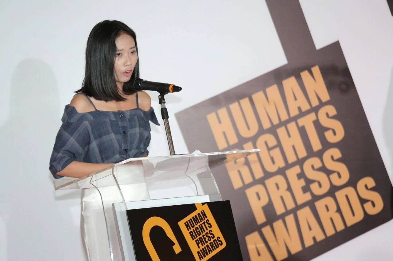 Eine Frau in einem schwarzen Kleid mit einem Dutt steht an einem Rednerpult mit einem Mikrofon und hält eine Rede vor einem Banner mit der Aufschrift 'Human Rights Press Awards'.