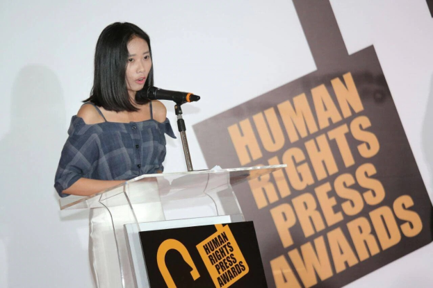 Eine Frau in einem schwarzen Kleid mit einem Dutt steht an einem Rednerpult mit einem Mikrofon und hält eine Rede vor einem Banner mit der Aufschrift 'Human Rights Press Awards'.