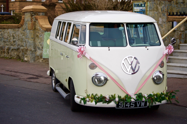 Ein weißer und pinker VW-Bus mit floralen Verzierungen, der vor einer Steinmauer geparkt ist, mit einer Treppe auf der rechten Seite und einem Gebäude im Hintergrund.