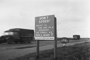 Schwarzes und weißes Foto eines Trucks, der auf einer Straße mit einem "Don't Speed"-Schild im Vordergrund und anderen Fahrzeugen, Gras, Pfählen und einem klaren Himmel im Hintergrund fährt.
