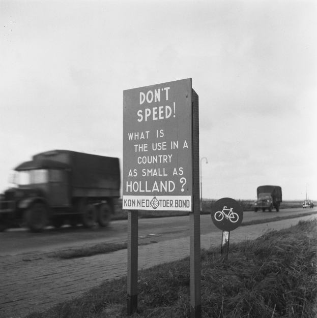 Schwarzes und weißes Foto eines Trucks, der auf einer Straße mit einem "Don't Speed"-Schild im Vordergrund und anderen Fahrzeugen, Gras, Pfählen und einem klaren Himmel im Hintergrund fährt.