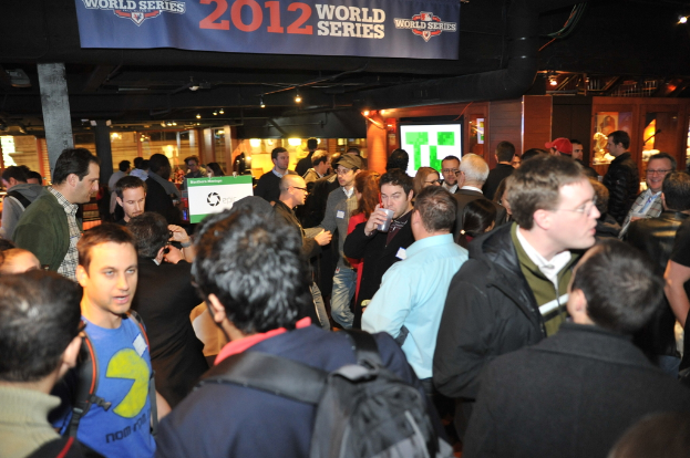 Eine große Menschenmenge bei einem Event mit einem Bildschirm, der ein Bild anzeigt, Lichtern und einer hölzernen Wand im Hintergrund.