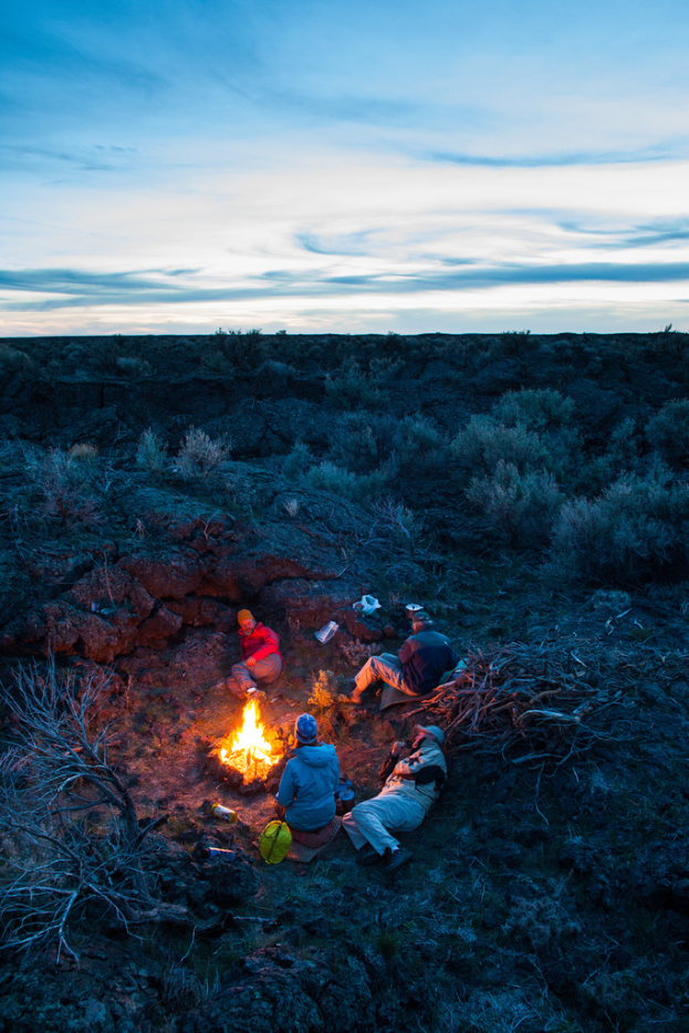 Ein Lagerfeuer mit Menschen, die darauf sitzen und liegen, mit Mützen tragend, mit einem Beutel und anderen Gegenständen auf dem Boden, umgeben von Bäumen und Himmel.