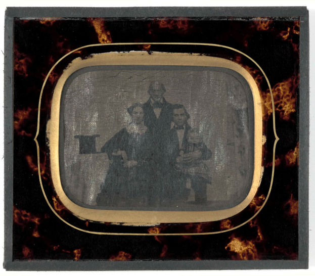 Ein altes Foto eines Mannes im Anzug und einer Frau in unbekannter Kleidung, die vor einem Fernseher stehen und ernst schauen.