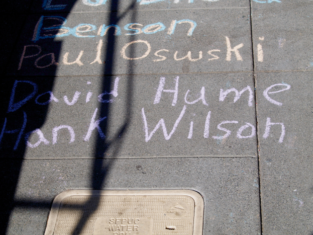 Ein Gehweg mit Kreide-Schrift, die wie ein Denkmal aussieht, ein Gullydeckel unten und ein Baumschatten auf der linken Seite.