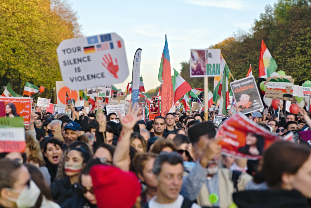 Eine große Gruppe von Menschen mit Schildern und Flaggen im Zusammenhang mit der iranischen Revolution im Hintergrund und einem klaren blauen Himmel.