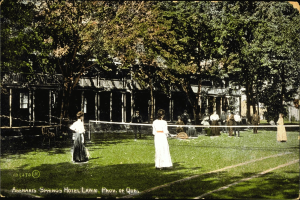 Schwarz-weiß-Foto einer Gruppe von Menschen, die Tennis auf einem Rasenplatz vor dem Abenakis Springs Hotel Lawn in Provo, Quebec spielen, mit einem Netz, Bäumen und einem Gebäude im Hintergrund.