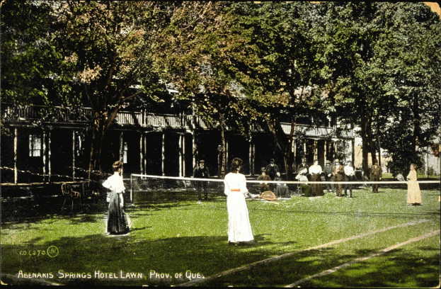 Schwarz-weiß-Foto einer Gruppe von Menschen, die Tennis auf einem Rasenplatz vor dem Abenakis Springs Hotel Lawn in Provo, Quebec spielen, mit einem Netz, Bäumen und einem Gebäude im Hintergrund.