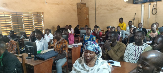 Eine Gruppe von Menschen sitzt an Tischen in einem classroom mit Monitoren, Taschen, Büchern, Papieren und anderen Gegenständen auf den Tischen, ein Fenster auf der linken Seite und Kartons und ein Fernseher an der Wand im Hintergrund, an einer Schulung zur Techniknutzung teil.