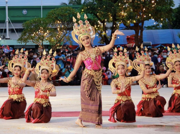 Eine Gruppe von Frauen in traditioneller Thai-Kleidung tanzt vor einem Publikum im Freien mit Bäumen im Hintergrund.