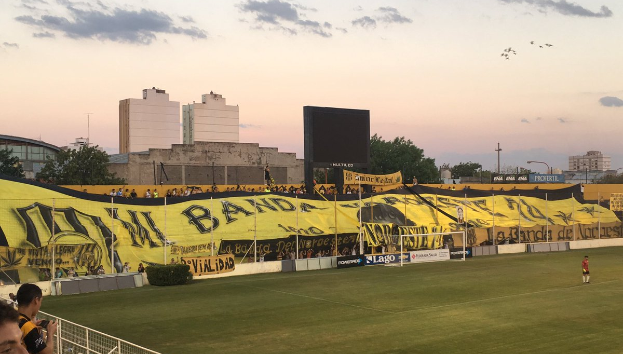 Ein Fußballfeld mit einer Gruppe von Menschen darauf, umgeben von einem Metallzaun, einem Torpfosten, einer Fahne mit der Aufschrift "Boca Juniors vs Real Sociedad", einigen Pflanzen, einem Bildschirm, einigen Pfählen, der Rinde eines Baumes, einigen Bäumen, einigen Gebäuden und einem bewölkten Himmel.