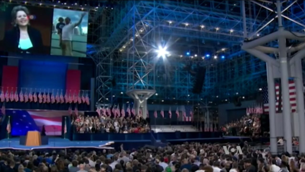 Eine große Menschenmenge sitzt vor einer Bühne mit einem Rednerpult, Mikrofon, Fahnen, einem Bildschirm und Beleuchtungsausrüstung und wartet auf eine Debatte.