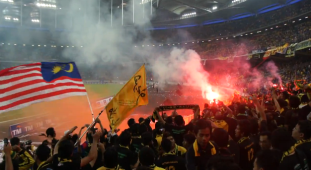 Eine große Menschenmenge in einem Stadion mit Bengalos, Fahnen und einer Kamera, mit Rauch und Schildern mit der Aufschrift "Malaysia vs Malaysia Live Streaming" im Hintergrund.