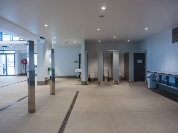 Großes öffentliches WC mit mehreren Toiletten, Säulen, einem Mülleimer, einer Tür, einem Schild, einem Feuerlöscher und Deckenleuchten.