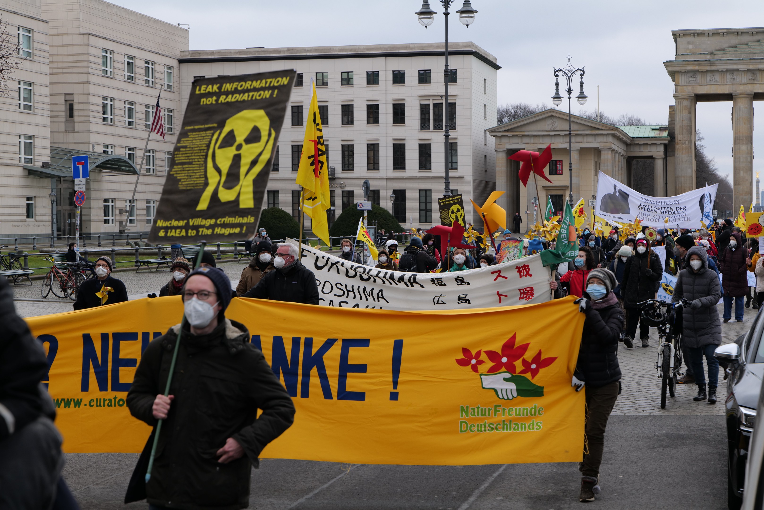 Demonstration gegen Atomkraft in Deutschland, mit einer großen Gruppe von Menschen, die Transparente und Fahnen schwenken und eine Straße entlangmarschieren, Fahrzeuge auf der rechten Seite und Gebäude, Bäume und ein Tor im Hintergrund.