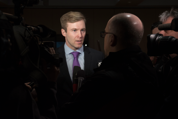 Ein Mann im Anzug und Krawatte, der eine Brille trägt, wird von Reportern interviewt, während zwei Personen mit Kameras rechts und links von ihm stehen und ein Mikrofon vor ihm gehalten wird.