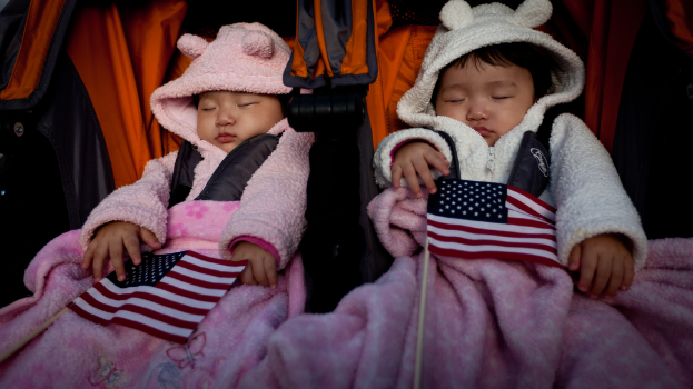 Zwei Babys schlafen in einem orange-schwarzen Babywagen mit weiß-roten-blauen Fahnen im Hintergrund und pinken Decken unten.