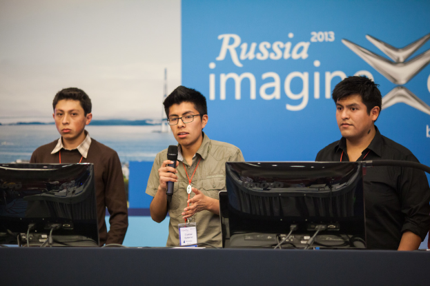 Drei Jungen stehen neben einem Schreibtisch mit zwei Computersystemen, einer spricht in ein Mikrofon, trägt ID-Karten mit roten Markierungen, vor einer Wand mit einer Werbetafel, auf der "Russia Imagine 2013" steht.