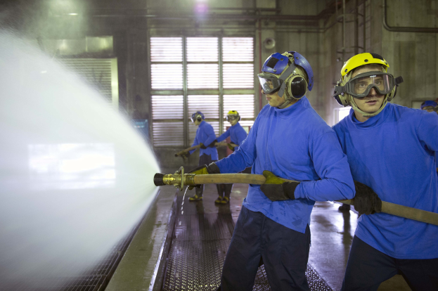 Gruppe von Männern in blauen Hemden und gelben Helmen arbeitet an Maschinen, wobei einer einen Schlauch hält und Wasser auf den Boden sprüht, in einer Fabrikumgebung mit sichtbaren Rohren, Fenstern und Lampen.