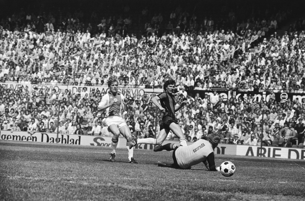Schwarzes Foto von Männern, die auf einem Feld Fussball spielen, mit Zuschauern im Hintergrund und einer "Bundesliga 1978-79 Ajax 1-1 Hannover 96"-Tafel.