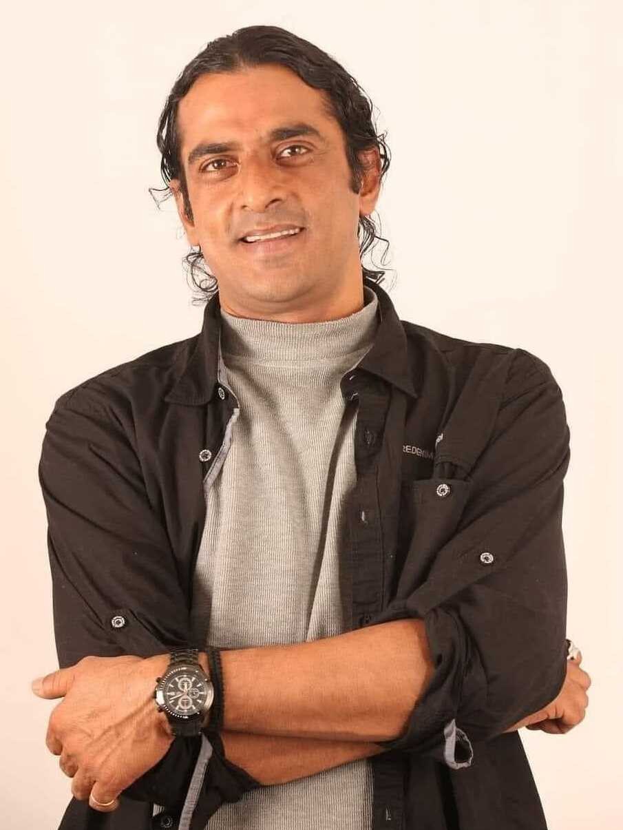 Ein Mann in einem schwarzen und aschfarbenen Kleid mit verschr├Ąnkten Armen, der eine Uhr tr├Ągt, vor einem wei├čen Hintergrund stehend und nachdenklich aussehend.