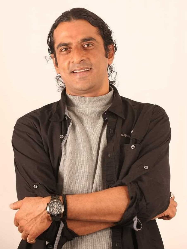 Ein Mann in einem schwarzen und aschfarbenen Kleid mit verschr├Ąnkten Armen, der eine Uhr tr├Ągt, vor einem wei├čen Hintergrund stehend und nachdenklich aussehend.