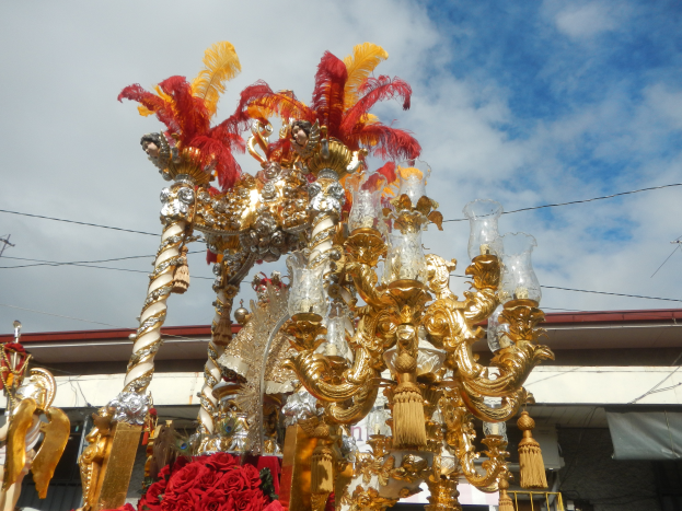 Ein prächtiger goldener und roter Wagen, geschmückt mit Blumen und anderen Dekorationen, bei einem Karnevalsumzug. Im Hintergrund sind ein Gebäude, Strommasten mit Drähten und ein bewölkter Himmel zu sehen.