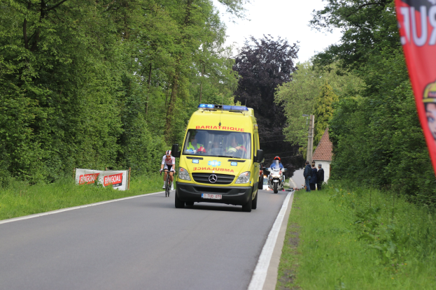 Ambulanz fährt auf einer Straße mit Fahrradfahrern daneben, umgeben von Gras, Bäumen, Häusern, Strommasten und einem klaren blauen Himmel.