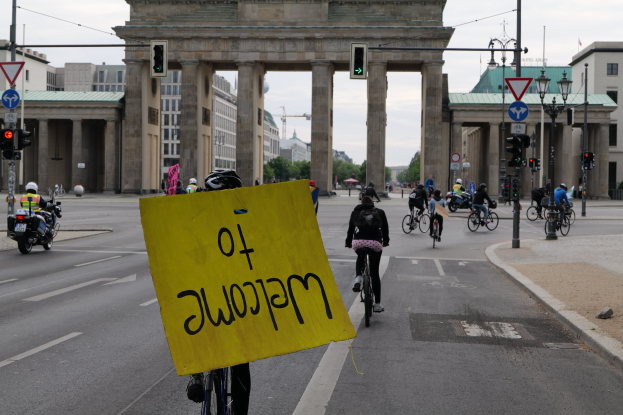 Gruppe von Radfahrern, die an der Brandenburgertor in Berlin vorbeifährt, einer hält ein gelbes Schild, mit Laternenpfählen, Verkehrsampeln, Gebäuden, Bäumen und einem klaren Himmel im Hintergrund.