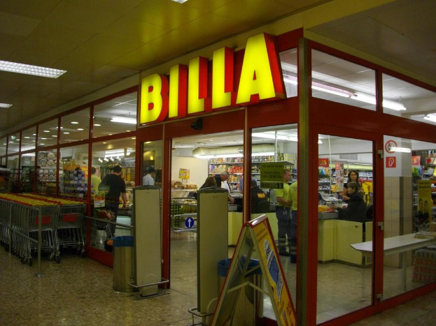 Ein Supermarkt mit einem Eingang in der Mitte, Mülltonnen daneben, eine Reihe von Einkaufswagen auf der linken Seite und einige Menschen drinnen, die unter der Deckenbeleuchtung Produkte durchstöbern.