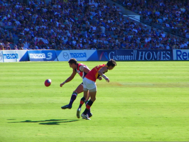 Zwei Personen in rot-weißer Kleidung stehen mit einem Ball in der Luft, während im Stadionhintergrund Banner und Publikum zu sehen sind.