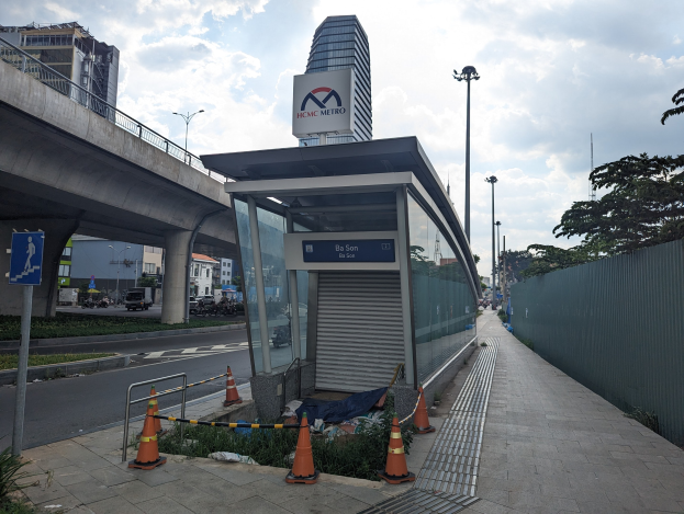 Busbahnhof mit Verkehrskegeln, Pflanzen und einem Schild neben einem hohen Gebäude, Fahrzeugen auf der Straße und einer Brücke unter einem bewölkten Himmel.