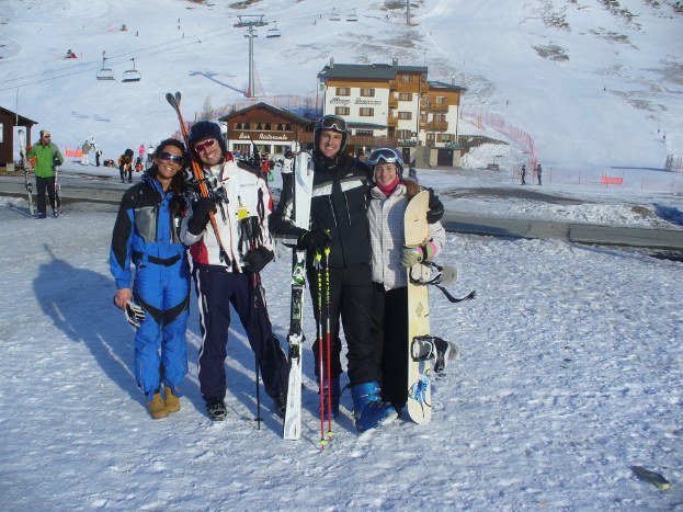 Vier Personen posieren für ein Foto mit Schlittschuhschutz und Werkzeugen vor einem Gebäude und anderen Menschen auf einem schneebedeckten Berg.