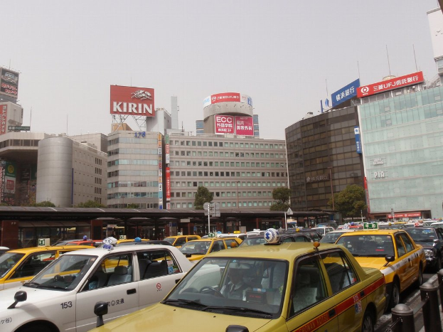 Eine Stadtansicht mit Fahrzeugen auf der Straße, Gebäuden, Bäumen und Masten mit Texttafeln, einem Zug im Hintergrund und einem Geländer unten rechts.