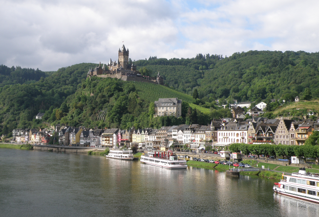Ein malerischer Blick auf den Rhein in Deutschland mit einer Burg auf einem Hügel im Hintergrund, Booten auf dem Fluss, Fahrzeugen auf einer näheren Straße und einem bewölktem Himmel.