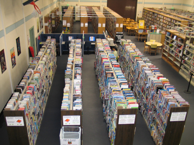 Ein Bibliotheksinneres mit Bücherregalen, Tischen und Stühlen vor einer Wand.