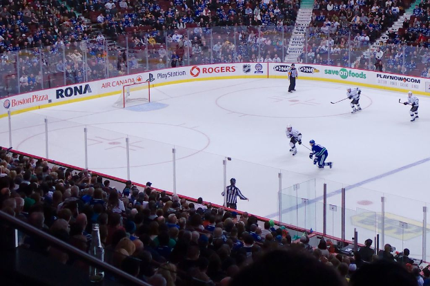 Vier Männer spielen Hockey auf einem Eisstadion, mit zwei in der Nähe des Tors und des Zauns stehenden, Zuschauern auf Stühlen im Vorder- und Hintergrund sowie einer Treppe im Hintergrund.