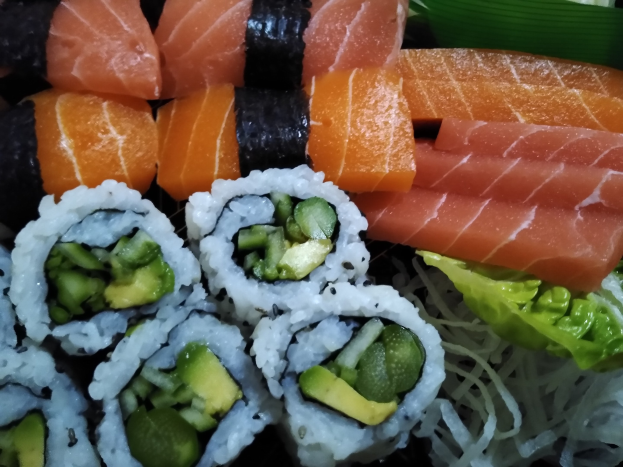 Ein schwarzer Teller mit verschiedenen Sushi-Stücken, darunter Lachs, Avocado und Gurke, umgeben von grünen Blättern.