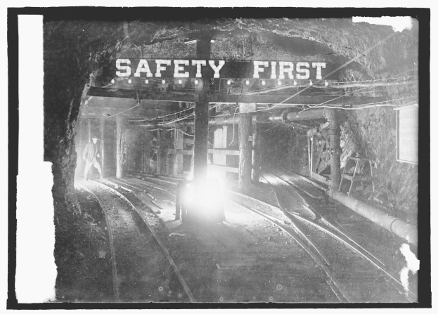 Schwarz-weißes Foto eines Zuges, der aus einem Tunnel fährt, mit einer Person auf den Schienen im Vordergrund, Hintergrund enthält Strommasten, Lampen und ein "Sicherheit geht vor"-Schild.