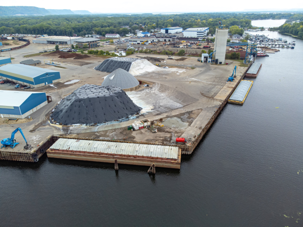 Luftaufnahme eines großen Gewässers mit einem zentralen Dock, umgeben von Gebäuden, Schuppen, Fahrzeugen, Kränen und anderen Strukturen, mit Bäumen, Hügeln und einem klaren blauen Himmel im Hintergrund.