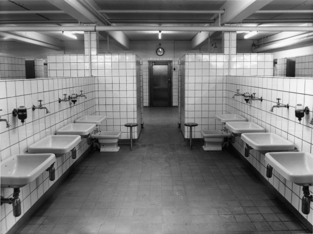 Ein Schwarz-Weiß-Foto einer öffentlichen Toilette mit Waschbecken, Wasserhähnen, Hockern, einer Wanduhr und Deckenlampen.
