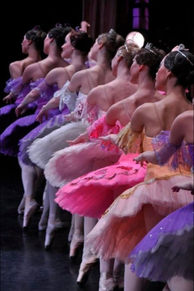 Balletttänzerinnen in rosafarbenen Tutus sitzen auf einer Bühne, beleuchtet von einem Scheinwerfer im Hintergrund, und führen synchronisierte Bewegungen aus.