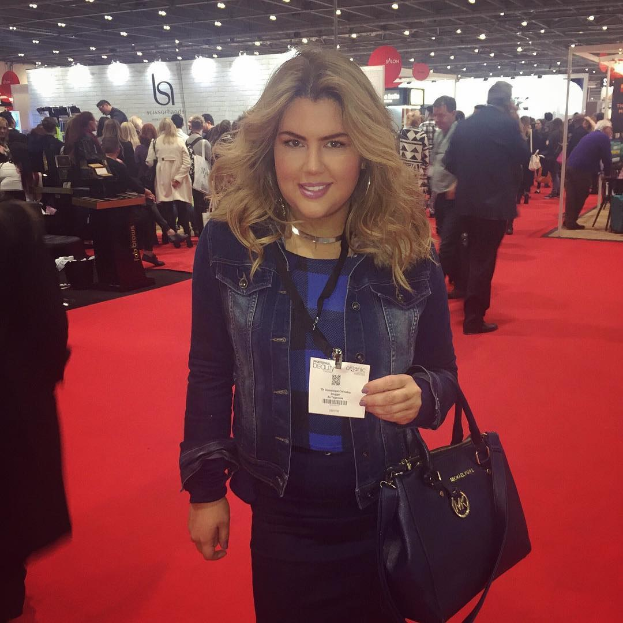 Frau auf einem roten Teppich an einer Modenschau, die lächelt und eine Handtasche hält, mit Menschen, Lichtern und Schildern im Hintergrund.