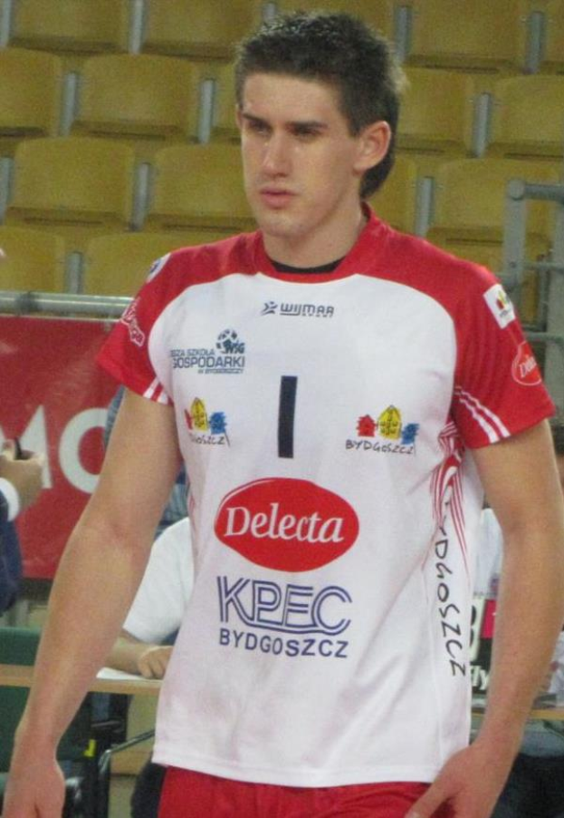 Ein Mann in einem roten und weißen Handballtrikot steht auf einem Feld, umgeben von Zuschauern, mit einem roten Banner im Hintergrund, auf dem 'Krasnoyarsk vs Bydgoszcz' steht.