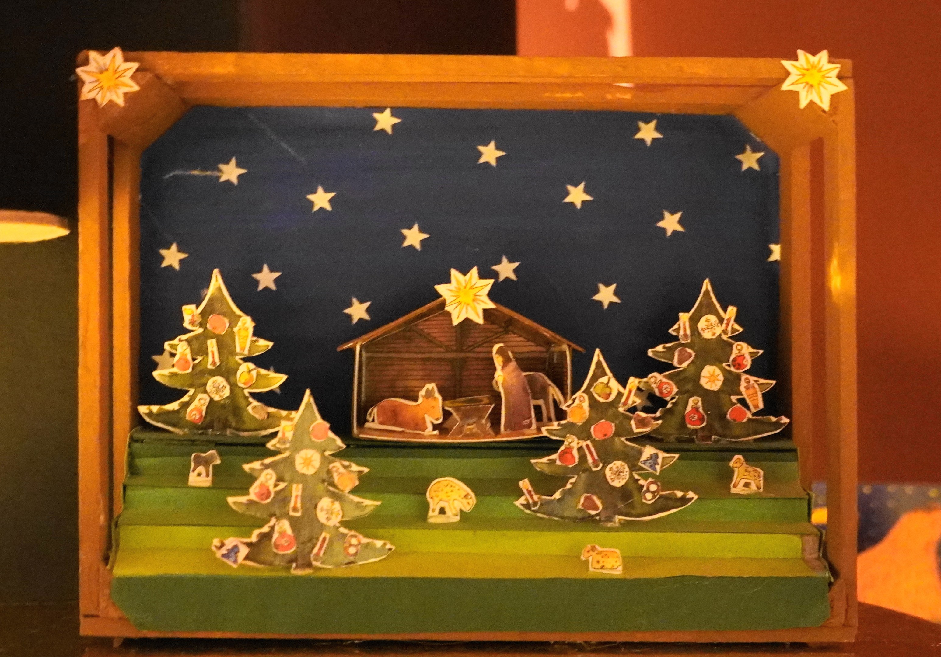 Eine Krippe in einer Holzkiste auf einem Tisch, dekoriert mit Weihnachtsbäumen, Tieren und einem Haus, mit einer Lampe auf der linken Seite und einer Wand im Hintergrund.