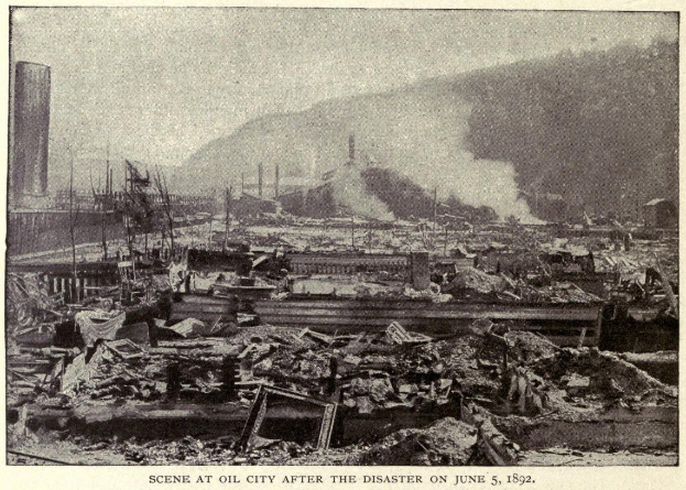 Schwarzes und weißes Foto einer Stadt nach einer Katastrophe am 5. Juni 1892, das Gebäude, Pfosten, Rauch, einen Hügel im Hintergrund und Text am Boden zeigt.