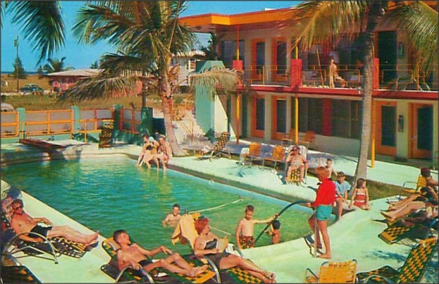 Ein Schwimmbad mit Menschen auf Stühlen im Vordergrund, Gebäuden und Bäumen im Hintergrund und Autos im Bild.