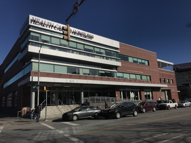 Großes Gebäude mit der Aufschrift "New York City Health Care Homeless Center" mit geparkten Autos, Passanten, Straßenmöbeln und Bäumen unter einem klaren Himmel.