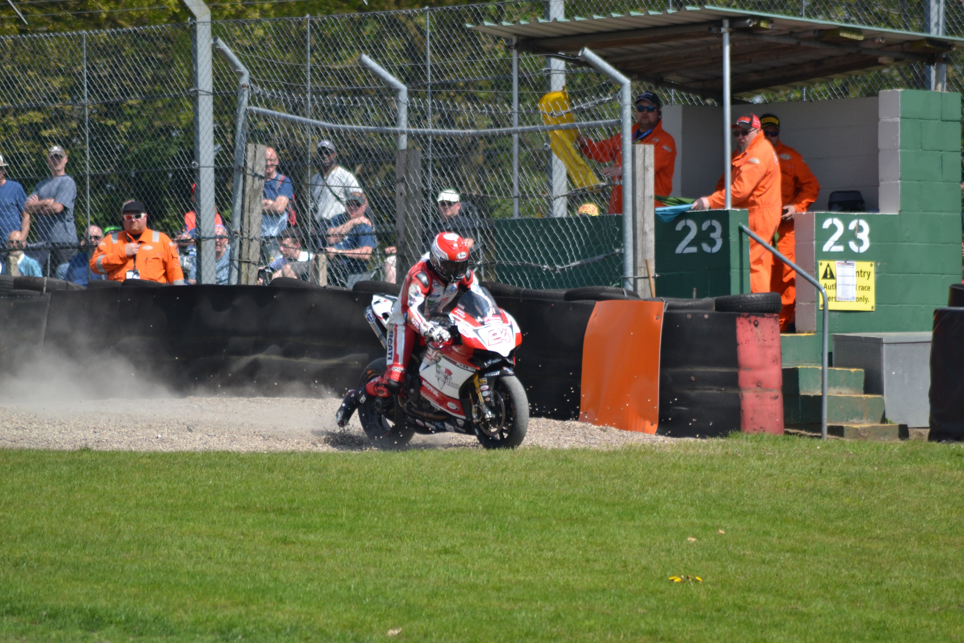 Ein Motorradfahrer in Helm und Handschuhen fährt über ein grasbewachsenes Feld, mit Zuschauern in Kappen und Schutzbrillen, einem eingezäunten Bereich, Treppen mit Geländern und einem nummerierten Schuppen im Hintergrund.
