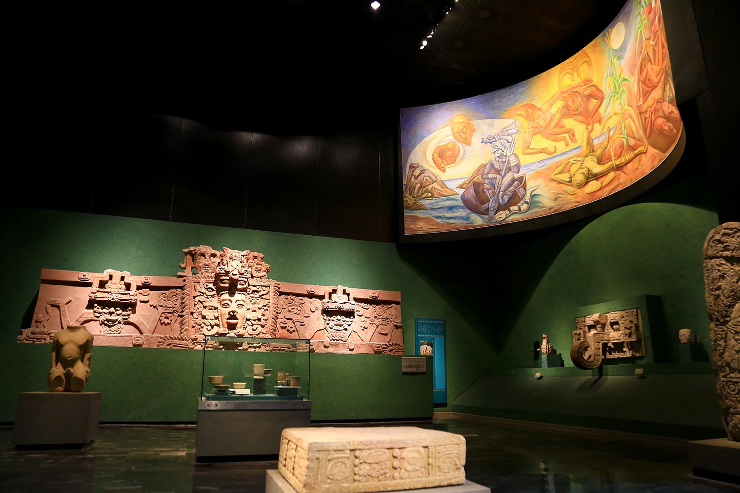 Ein Museumsraum mit verschiedenen Artefakten, darunter Skulpturen, Tischobjekte, eine Texttafel, eine Wandmalerei und Deckenleuchten im Mexiko-Stadt-Museum der Anthropologie.